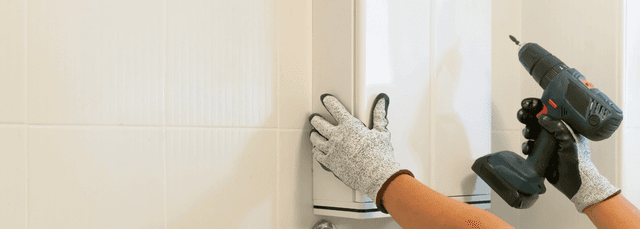 Hands wearing work gloves using cordless drill to install cabinet hardware on white kitchen cabinets