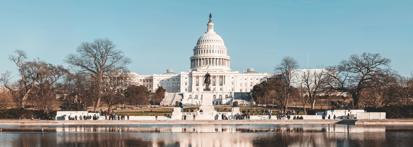 US Capitol building with its iconic dome reflected in water, surrounded by bare winter trees under a clear sky